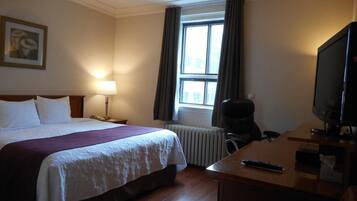 A hotel room with a bed, a desk with a television, a chair, a lamp, and a window with curtains.