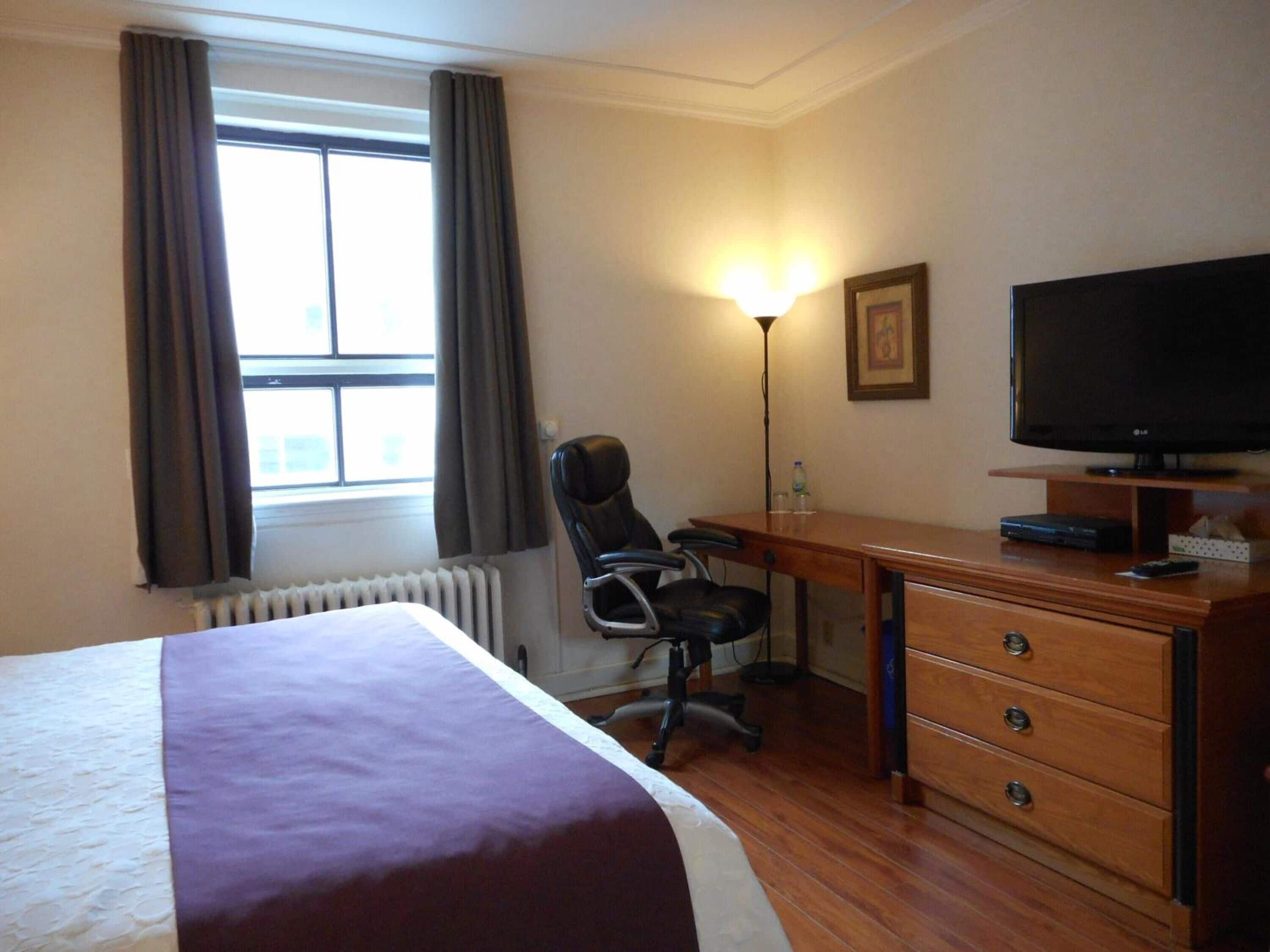 Una habitación de hotel con cama, escritorio, silla, televisor y radiador.