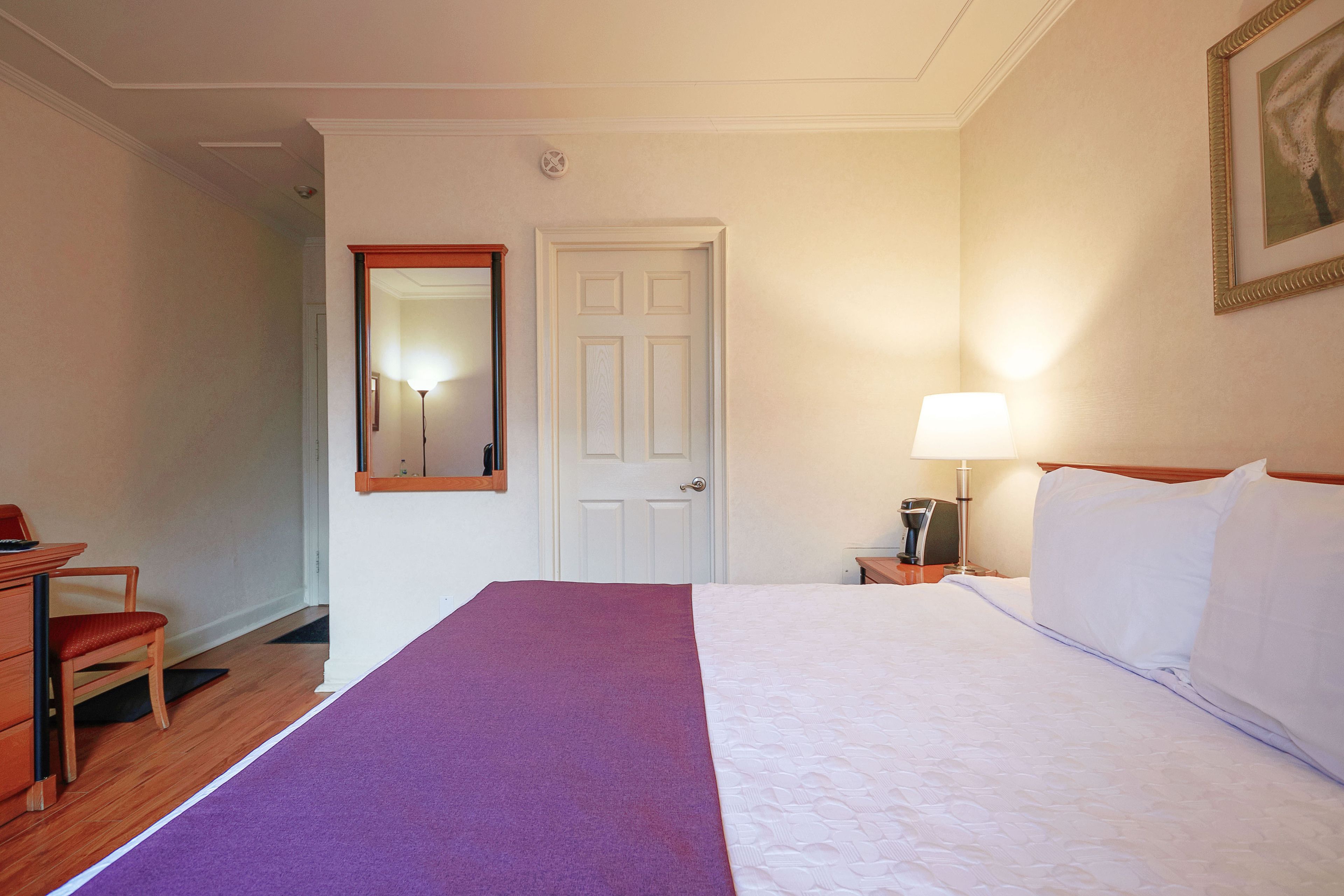 A hotel room with a bed, a desk, a chair, a door, and a nightstand with a lamp.