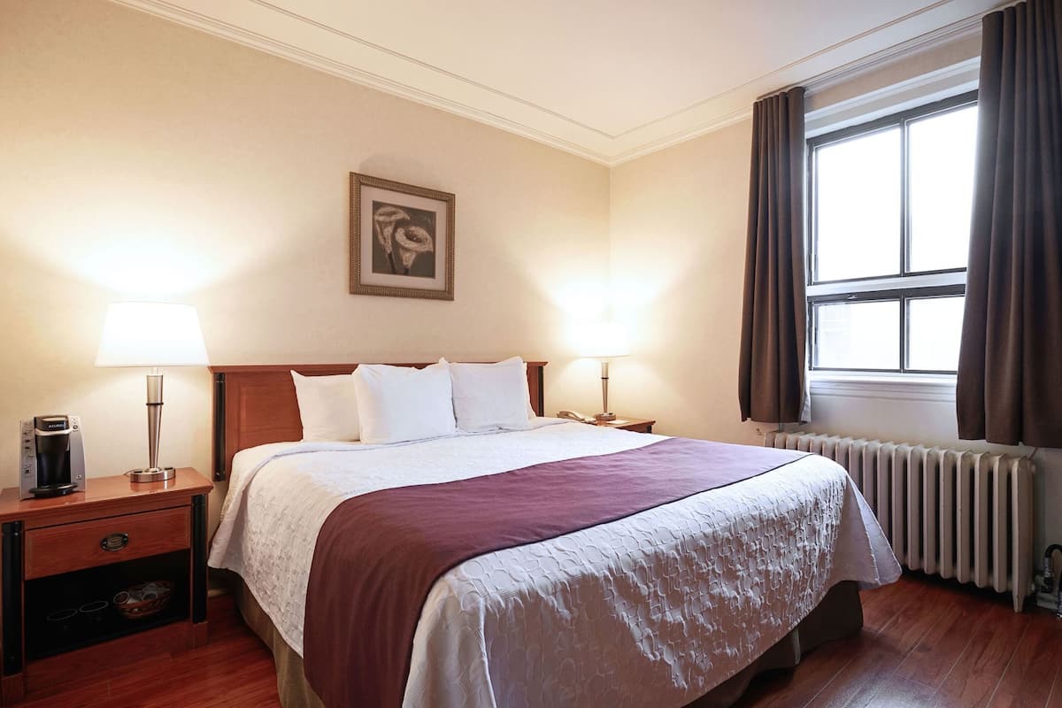 A hotel room with a bed, two bedside lamps, a coffee maker, a framed picture on the wall, and a window with curtains.