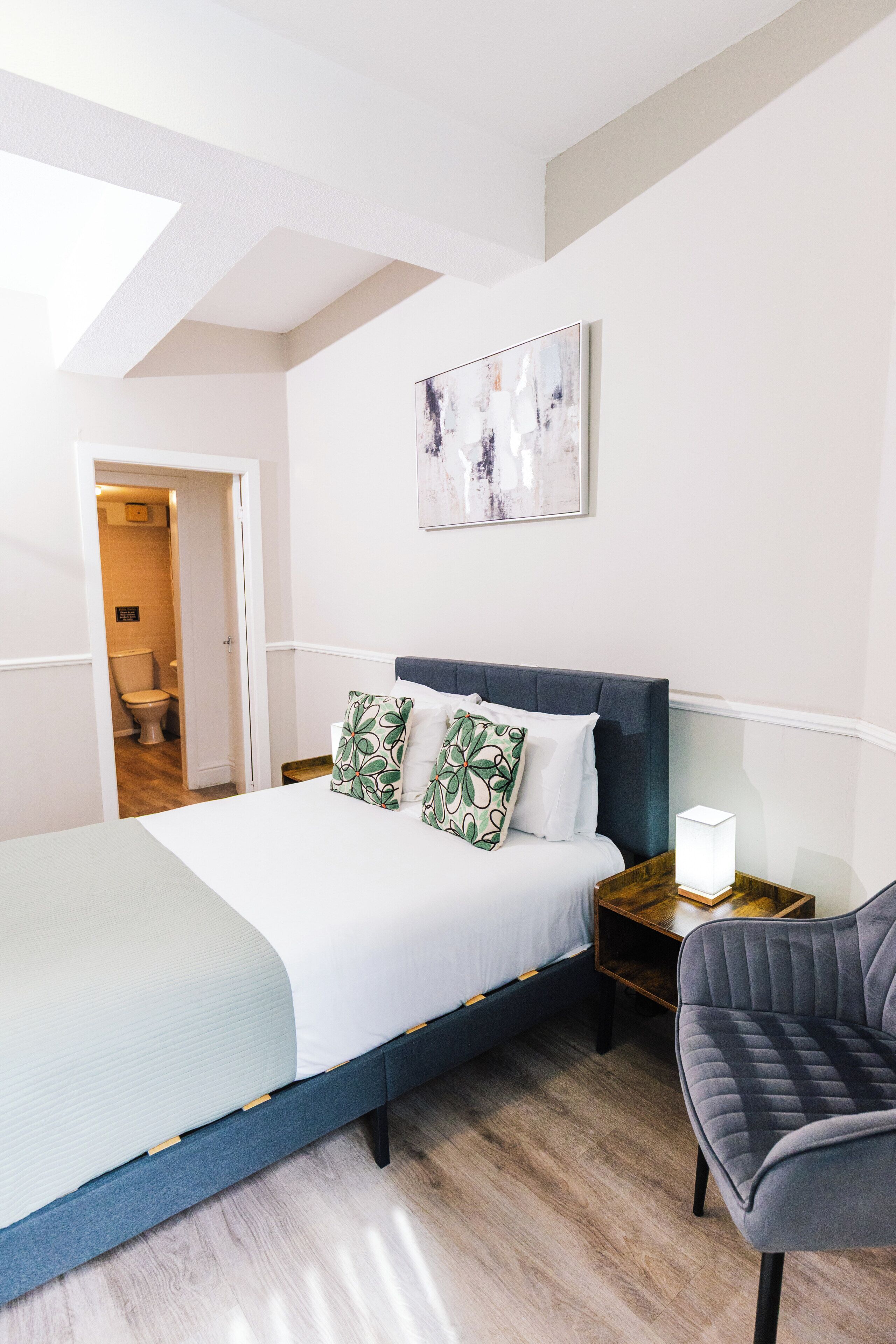 Un dormitorio moderno con una cama, una silla, una mesita de noche y un cuadro en la pared.