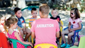 Un groupe d’enfants assis sur des chaises en plastique de couleurs vives, à l’extérieur, avec un homme portant un t-shirt rose sur lequel est inscrit le mot « ANIMATION » au premier plan.