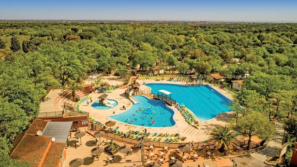 Un complexe hôtelier doté d’une grande piscine, de chaises longues et d’un espace de jeux pour enfants, le tout entouré d’arbres.