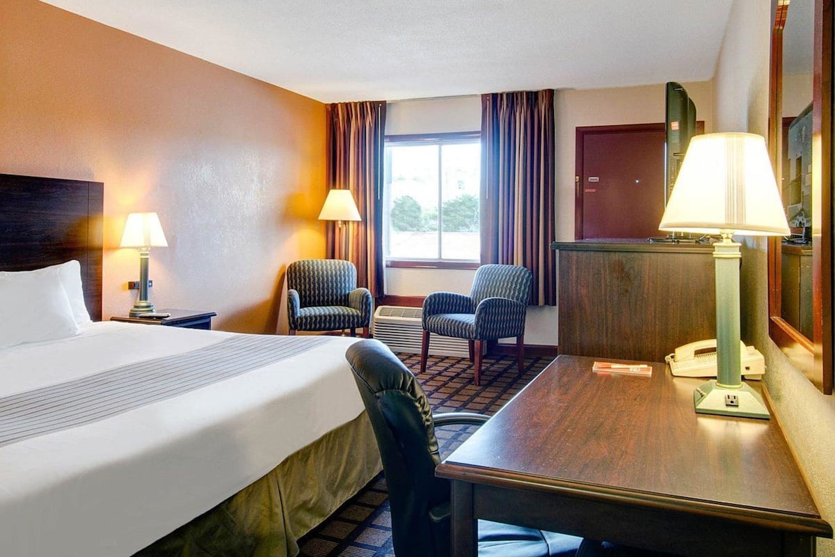 Une chambre d’hôtel comprenant un lit, deux fauteuils, un bureau avec une lampe, un téléphone et une fenêtre avec des rideaux.
