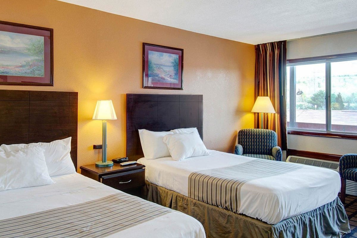 Une chambre d’hôtel avec deux lits, une table de chevet avec une lampe, une chaise et une fenêtre donnant sur un beau paysage.