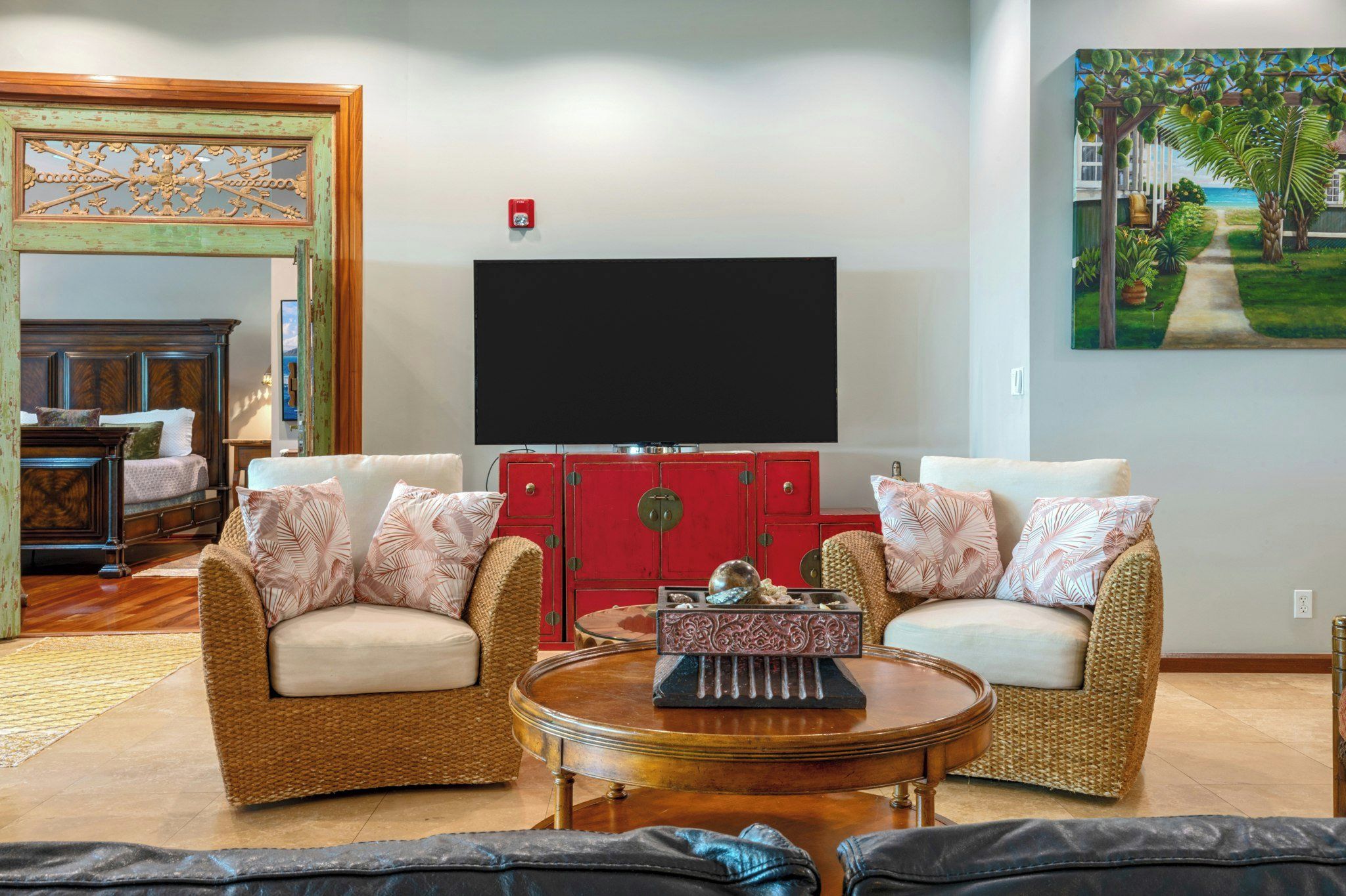 A living room with wicker furniture, a red cabinet, a flat-screen TV, and a painting of a tropical scene.