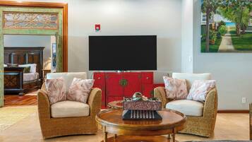 Una sala de estar con mobiliario de mimbre, un aparador rojo, un televisor de pantalla plana y un cuadro de una escena tropical.