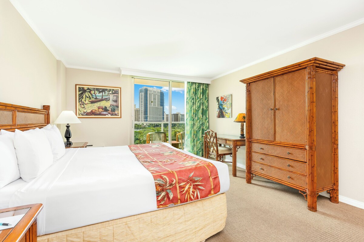 A hotel room with a large bed, a desk, a wardrobe, and a window with a city view.