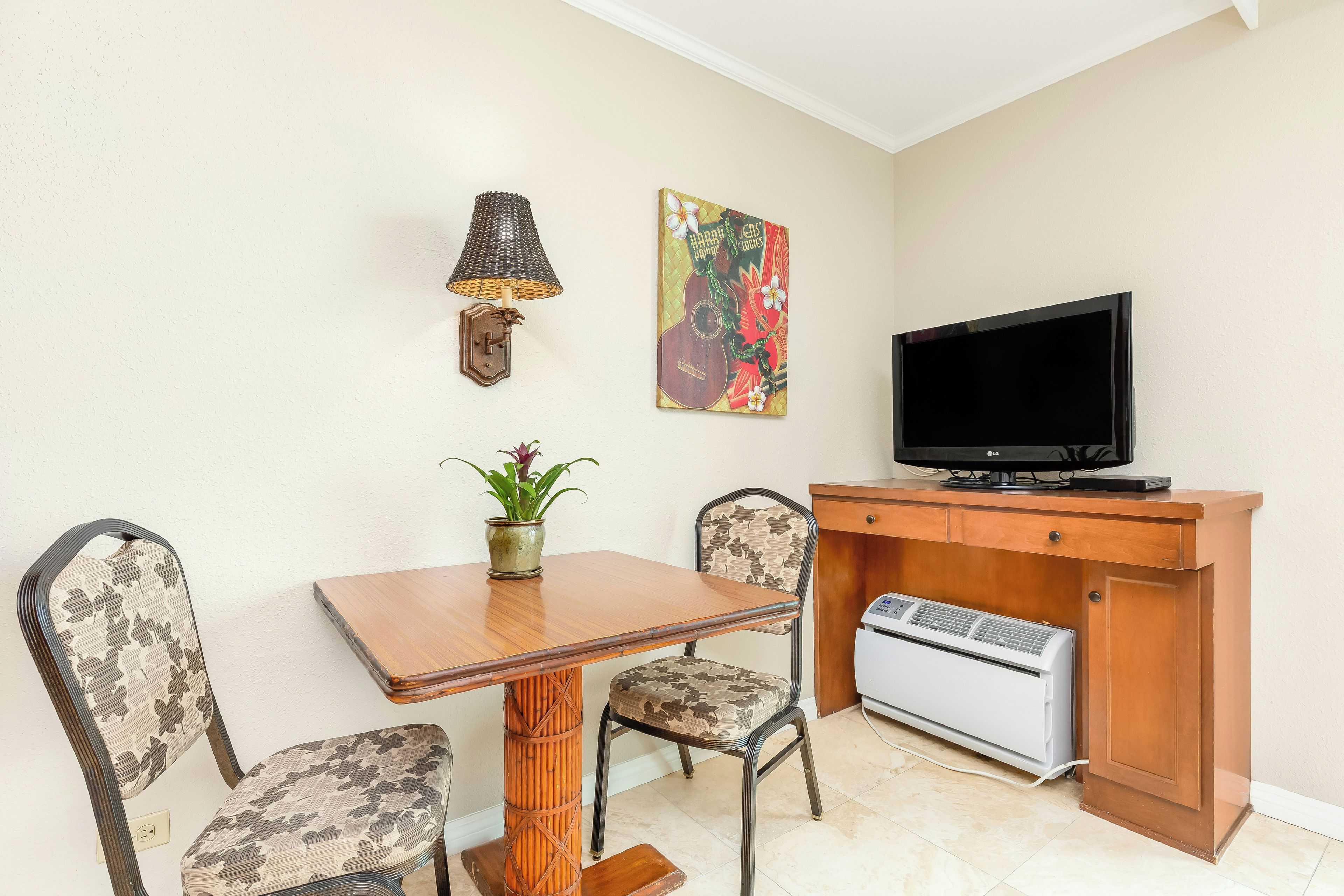 A small living area with a wooden table, two patterned chairs, a television, a wall-mounted lamp, a decorative painting, and a small heater.