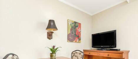 A small living area with a wooden table, two patterned chairs, a television, a wall-mounted lamp, a decorative painting, and a small heater.