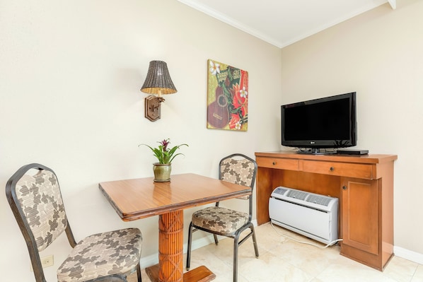 A small living area with a wooden table, two patterned chairs, a television, a wall-mounted lamp, a decorative painting, and a small heater.