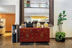 A hotel reception area with a coffee machine, a water dispenser, a red wooden cabinet with drawers, and a large mirror above the counter.