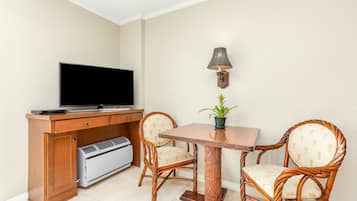 Een kamer met een televisie, een bureau met een printer, twee rieten stoelen, een kleine tafel en een wandlamp.