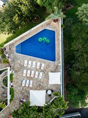 Vista aérea de una piscina con solárium de piedra, rodeada de sillones de playa y sombrillas, con árboles y vegetación al fondo.