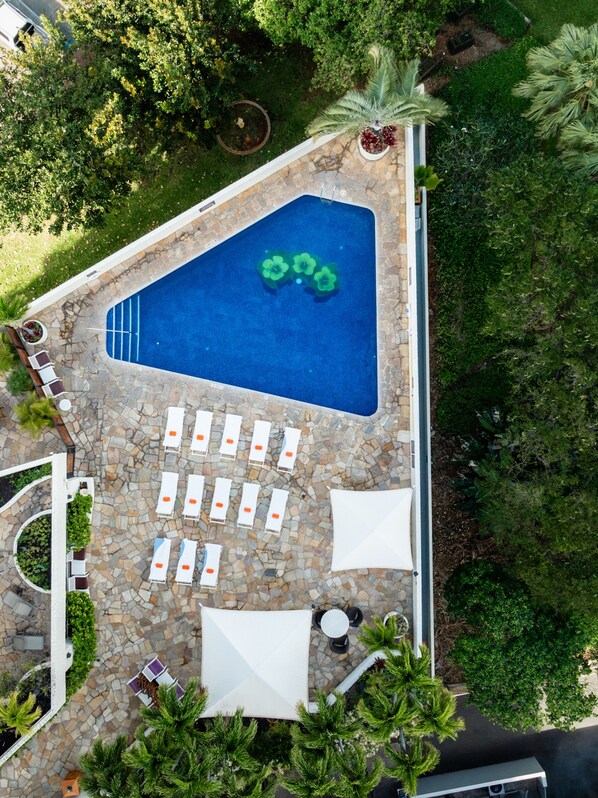 Vista aérea de una piscina con solárium de piedra, rodeada de sillones de playa y sombrillas, con árboles y vegetación al fondo.