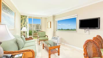 A hotel room with a sofa, armchairs, a flat-screen TV, a window with a view of buildings, and a wall-mounted air conditioning unit.
