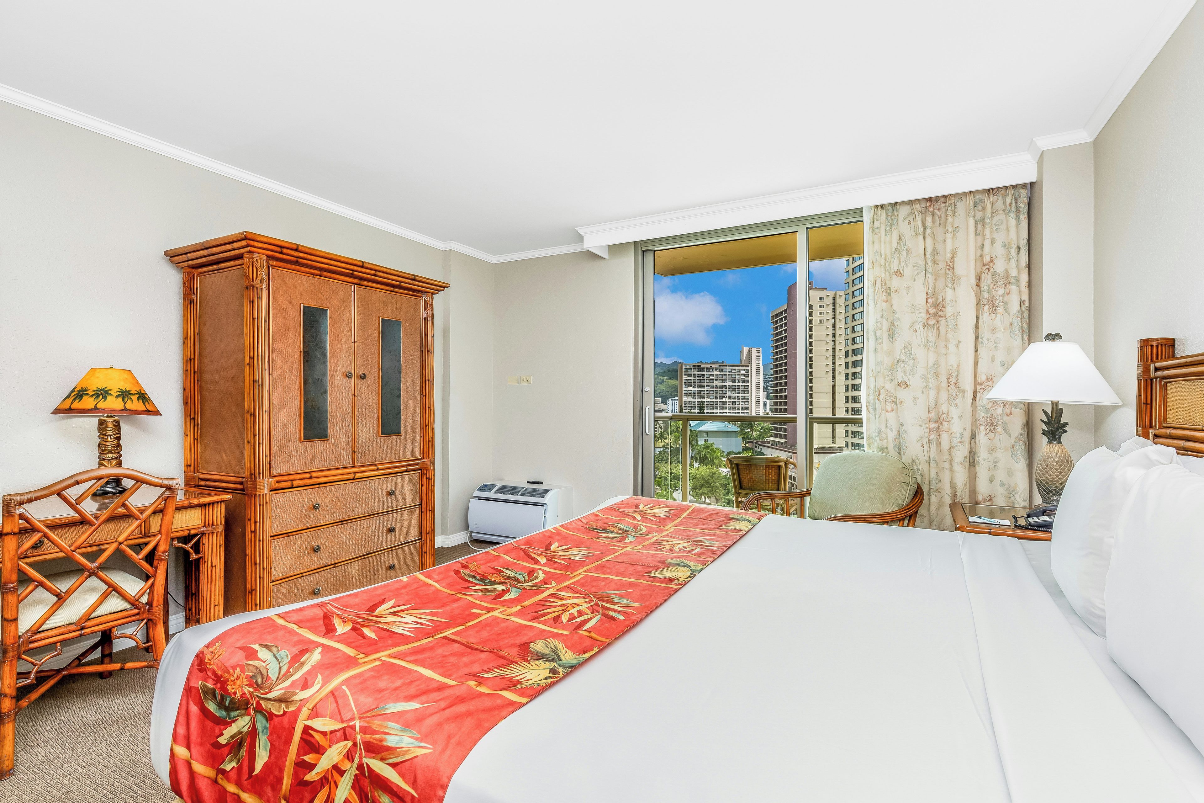 A hotel room with a bed, a desk, a wardrobe, a window with a city view, and a ceiling fan.