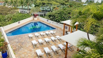 A swimming pool with lounge chairs, surrounded by greenery and tall buildings in the background.