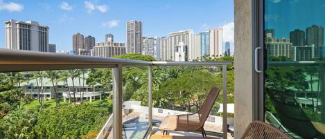 A balcony with a wooden table and chairs, overlooking a cityscape and lush greenery.