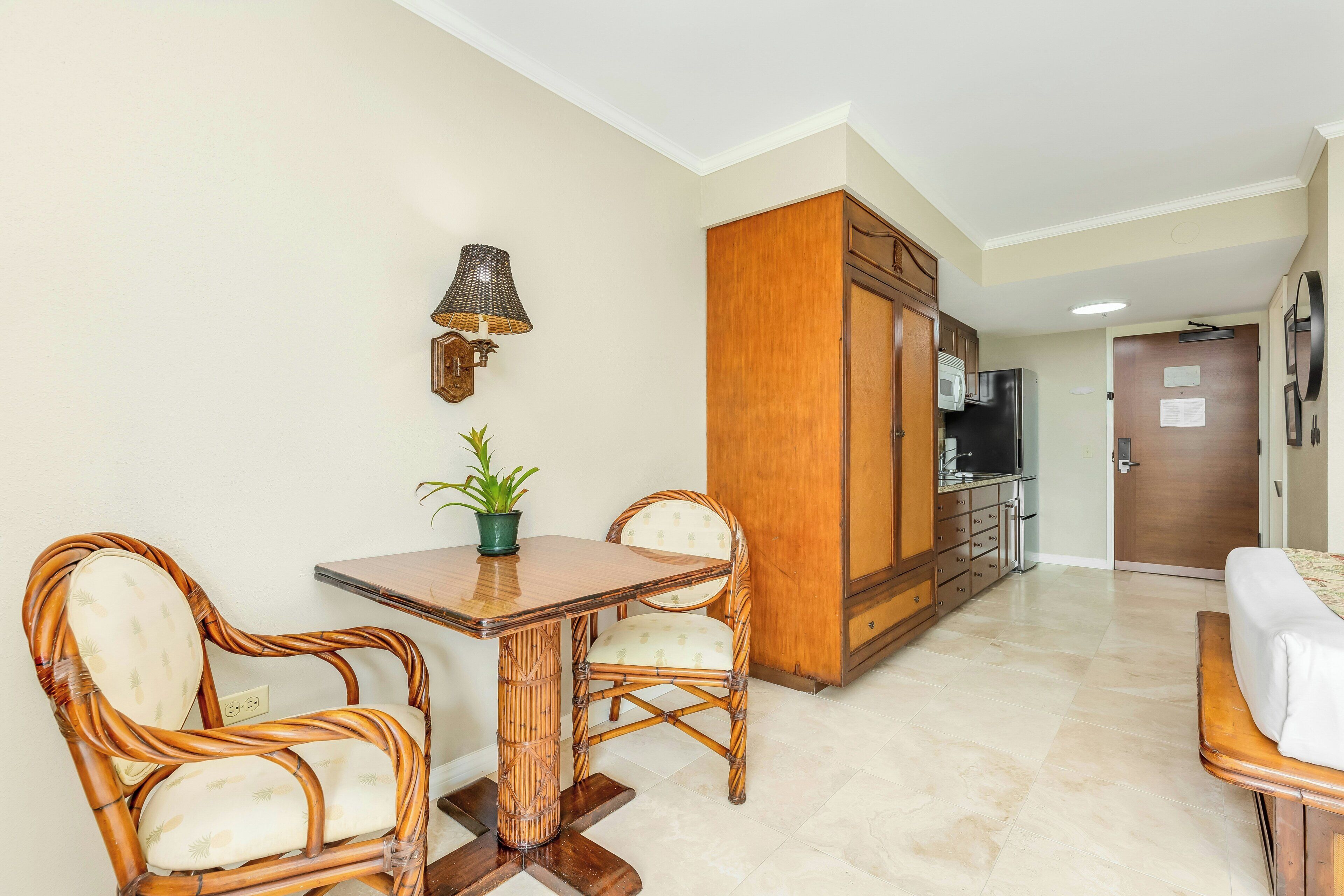 A room with a dining table, two chairs, a potted plant, a wardrobe, and a kitchenette.
