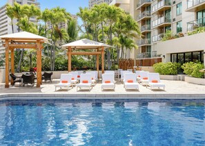 A poolside area with lounge chairs and a pergola, surrounded by palm trees and a building with balconies. - Luana Waikiki Hotel & Suites (Honolulu)