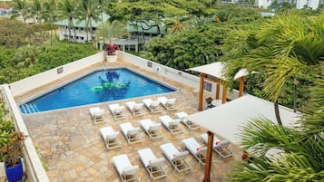 Piscina com espreguiçadeiras e área de estar coberta, cercada por vegetação e edifícios altos ao fundo.