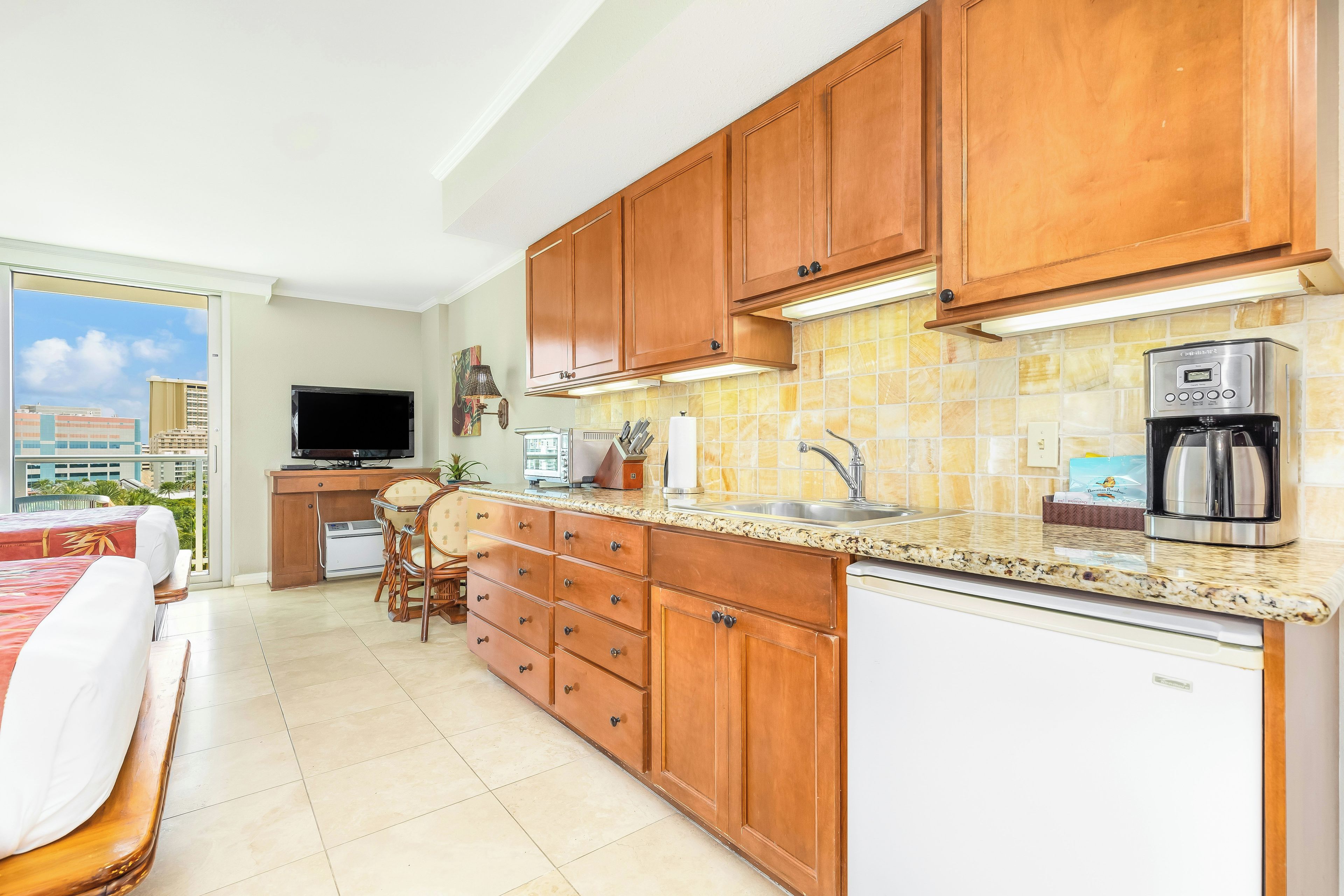 A modern kitchen with wooden cabinets, a built-in oven, a coffee maker, and a dining area with a TV and a plant.