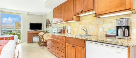 A modern kitchen with wooden cabinets, a built-in oven, a coffee maker, and a dining area with a TV and a plant.
