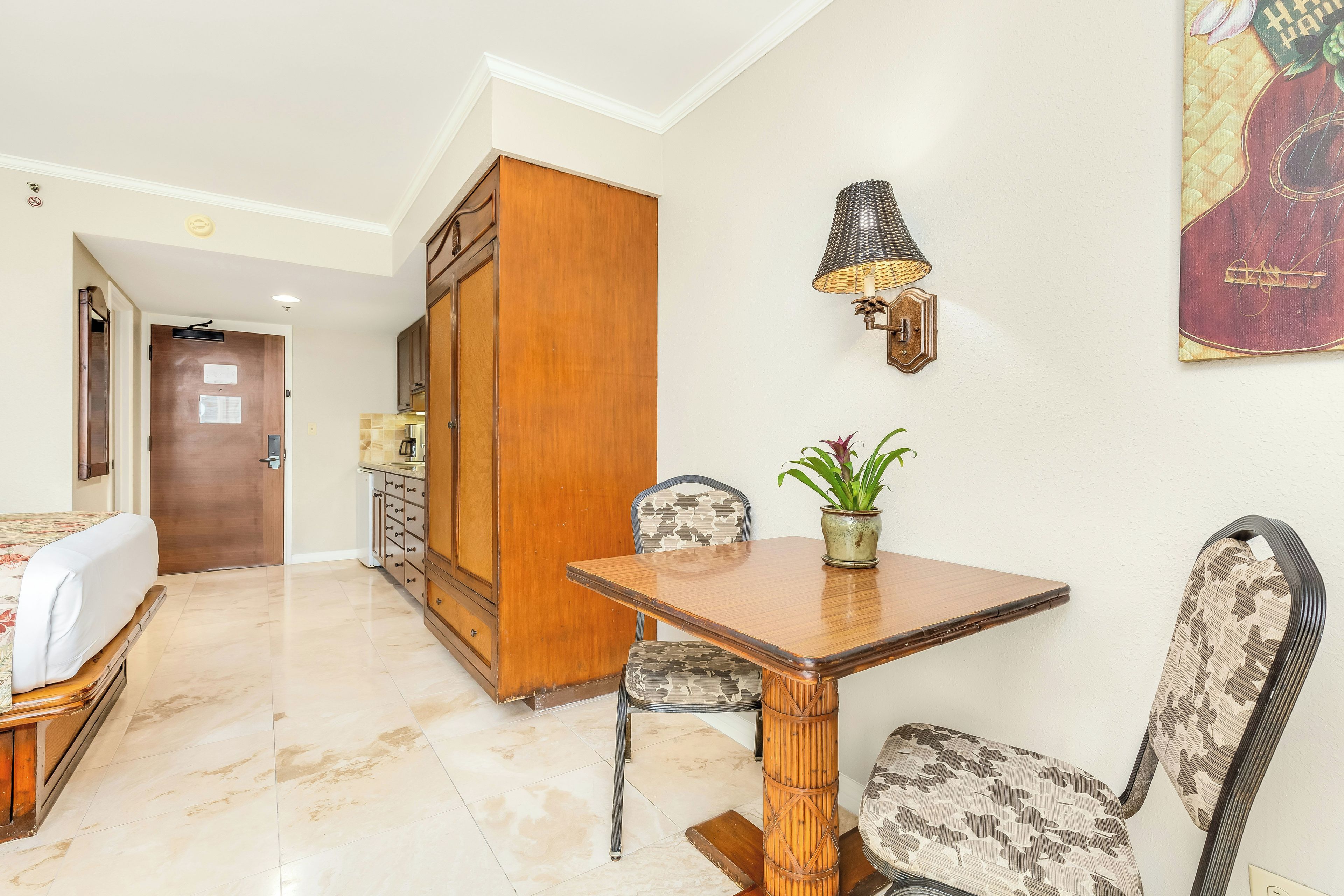 Quarto de hotel com mesa de jantar de madeira, cadeiras, planta em vaso, luminária na parede e cama com colcha florida.