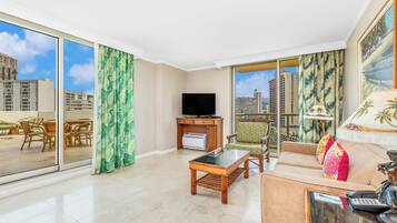 A spacious living room with a large window, a flat-screen TV, a coffee table, and a sofa with a decorative pillow.