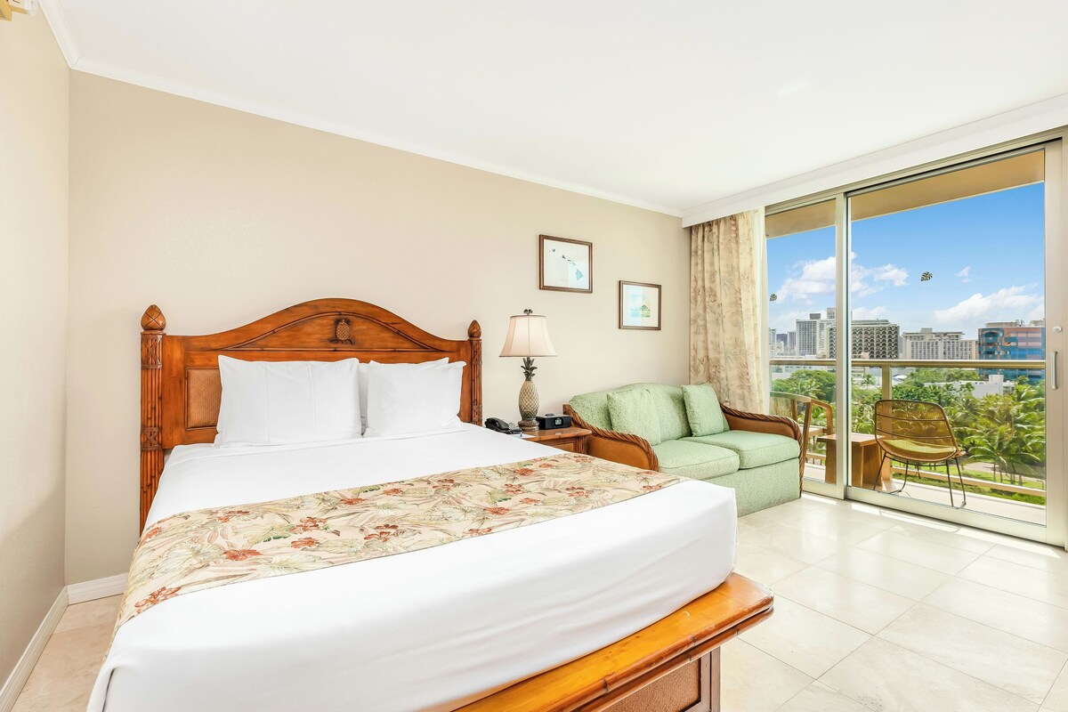 A hotel room with a large bed, a green sofa, a chair, a nightstand, a lamp, and a view of the city through the window.