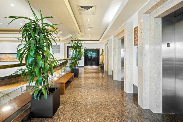 A hotel lobby with a marble floor, potted plants, a reception desk, and an elevator.