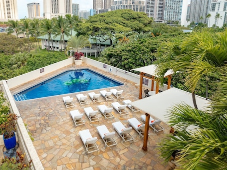 A swimming pool with lounge chairs, surrounded by greenery and tall buildings in the background.