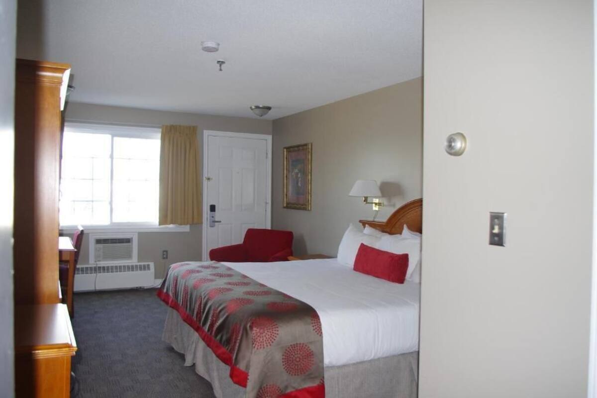 Quarto de hotel com cama, escrivaninha, cadeira, janela com cortinas, porta e luminária na parede.