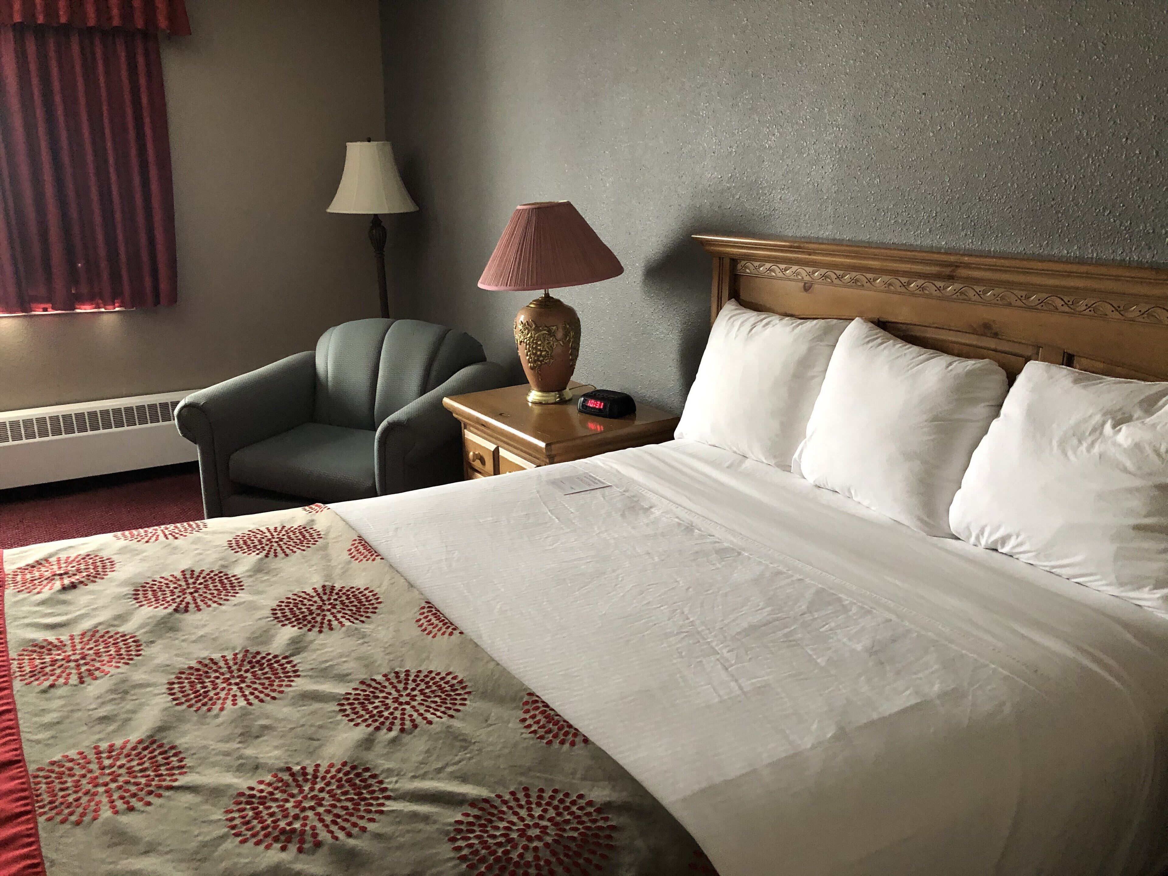 A hotel room with a bed, a chair, a nightstand, a lamp, and a heater.