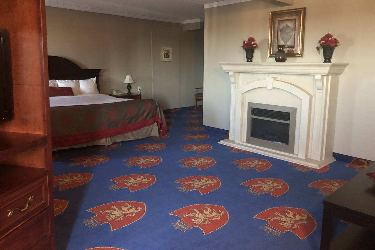 Quarto de hotel com cama, lareira, tapete com padrão vermelho e dourado, e cômoda de madeira.