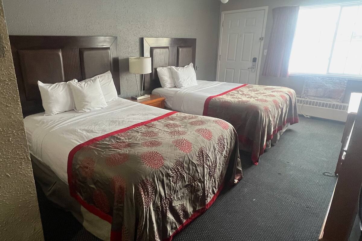Quarto de hotel com duas camas, cada uma com cabeceira, mesa de cabeceira, luminária e janela com cortinas.