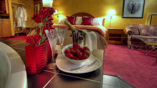 A hotel room with a bed, a bedside table with a lamp, a floral sofa, a dining table with a bowl of strawberries and champagne, and a red vase with roses. - South Hill Lodge (Prince Albert)