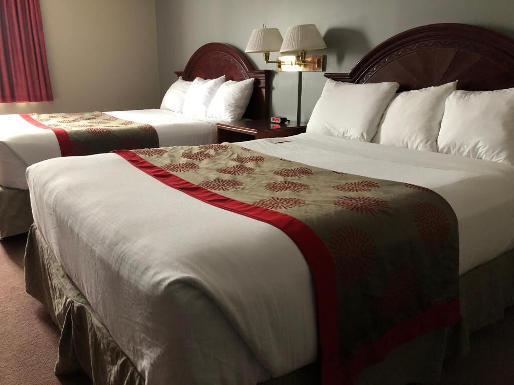 Une chambre d’hôtel avec deux lits, chacun agrémenté de draps blancs et d’une bordure décorative rouge. Il y a deux tables de chevet avec des lampes, ainsi qu’une lampe fixée au mur.
