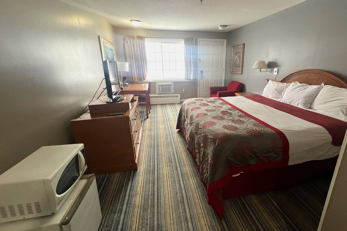 Quarto de hotel com cama, escrivaninha com computador, cadeira, micro-ondas e janela com cortinas.