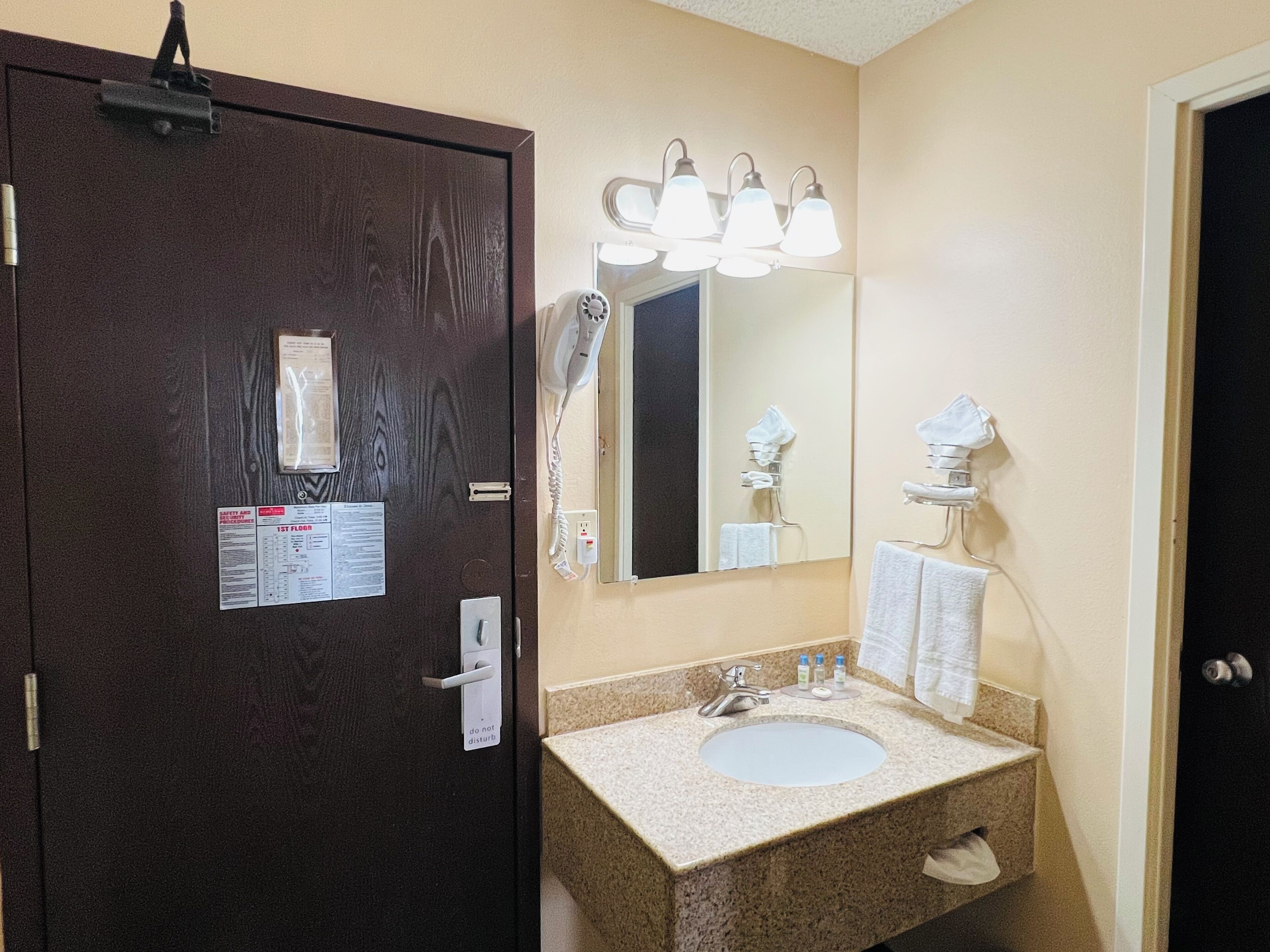 Un baño con puerta de madera oscura, lavamanos blanco, espejo con luces y un perchero para toallas.