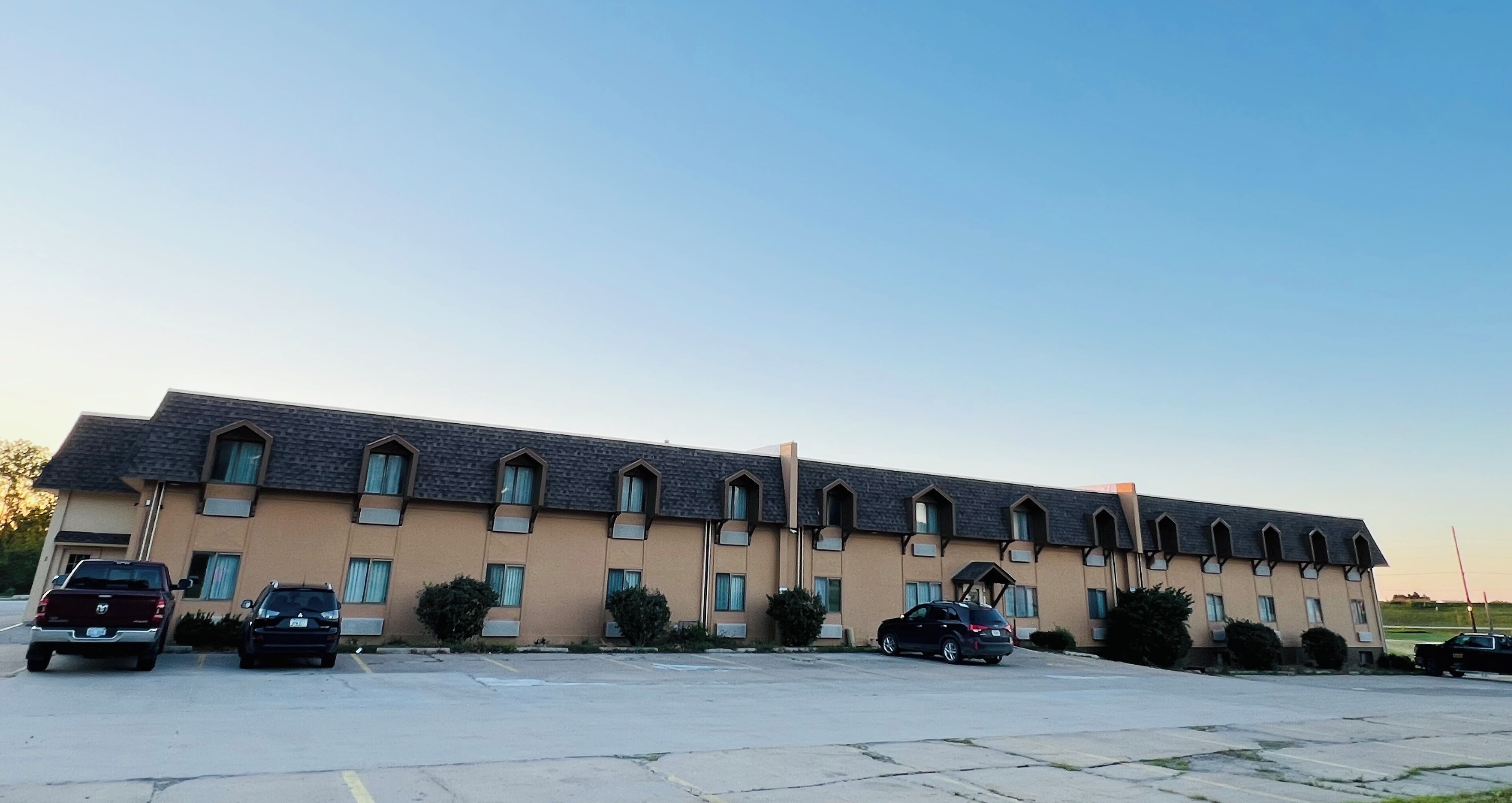Un edificio largo y de color beige con techo plano y numerosas ventanas, con estacionamiento al frente.