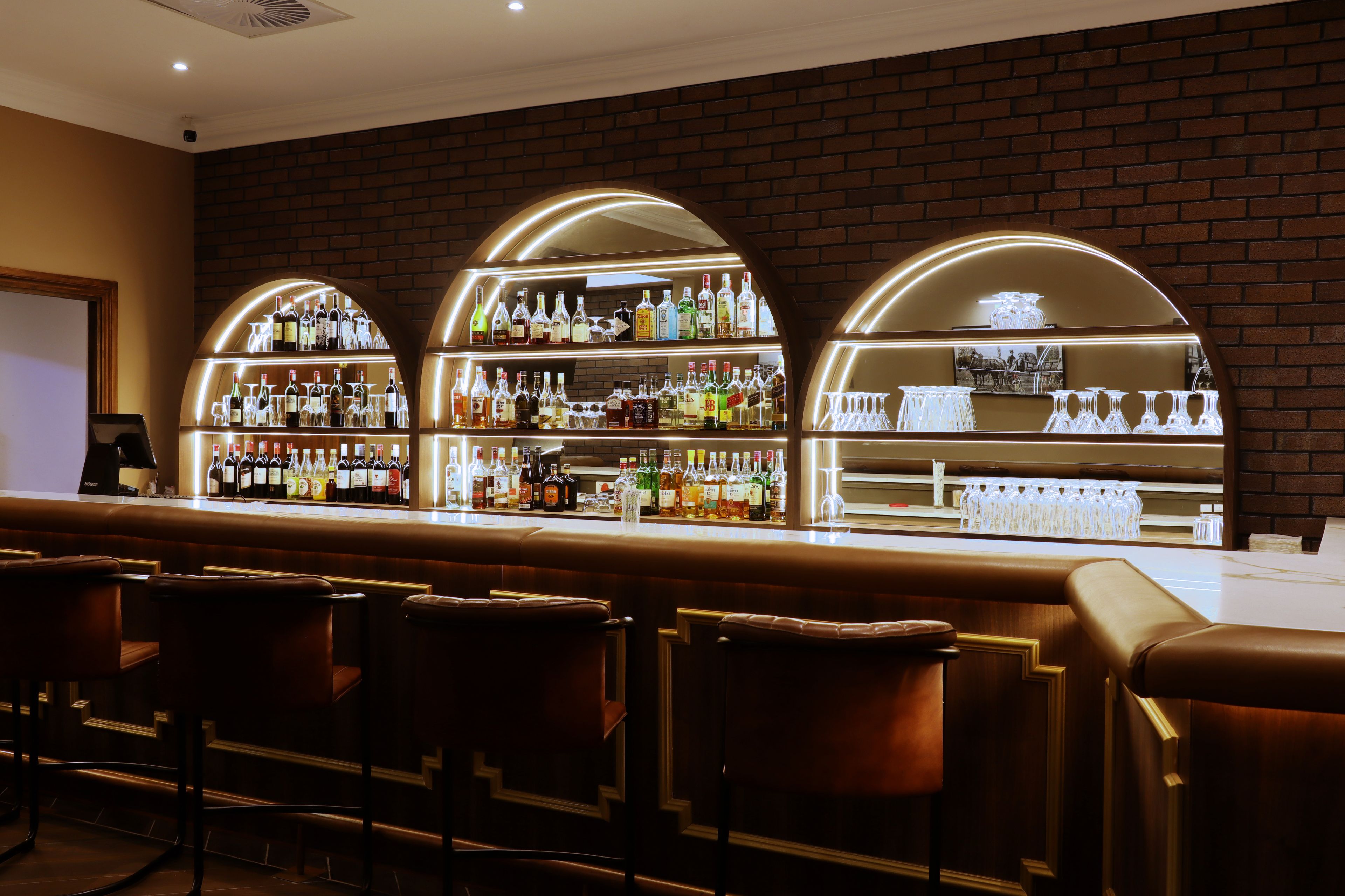 A bar area with multiple shelves of bottles and glasses, a seating area with chairs, and a brick wall backdrop.