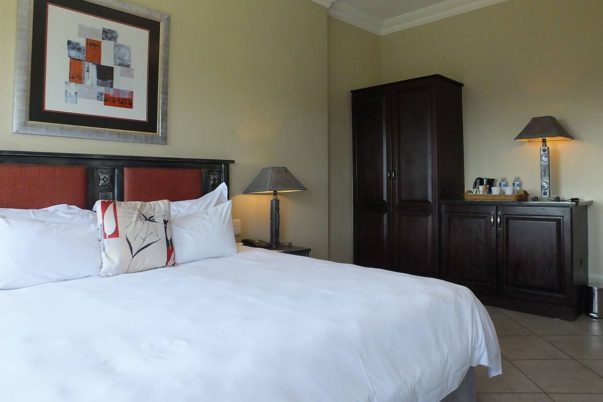 A hotel room with a large bed, a nightstand with a lamp, a wardrobe, and a framed artwork on the wall.