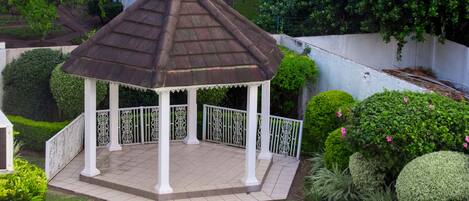A gazebo with a tiled roof and white columns, surrounded by well-maintained grass and hedges.