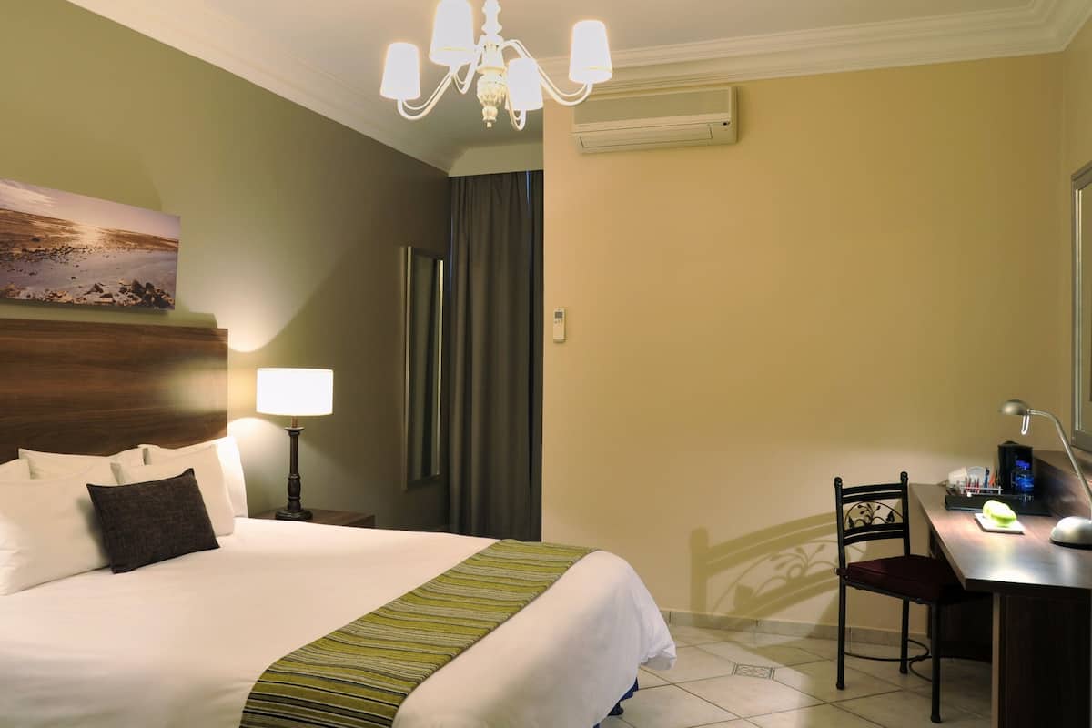 A hotel room with a bed, bedside lamps, a desk with a chair, a painting on the wall, and a chandelier.