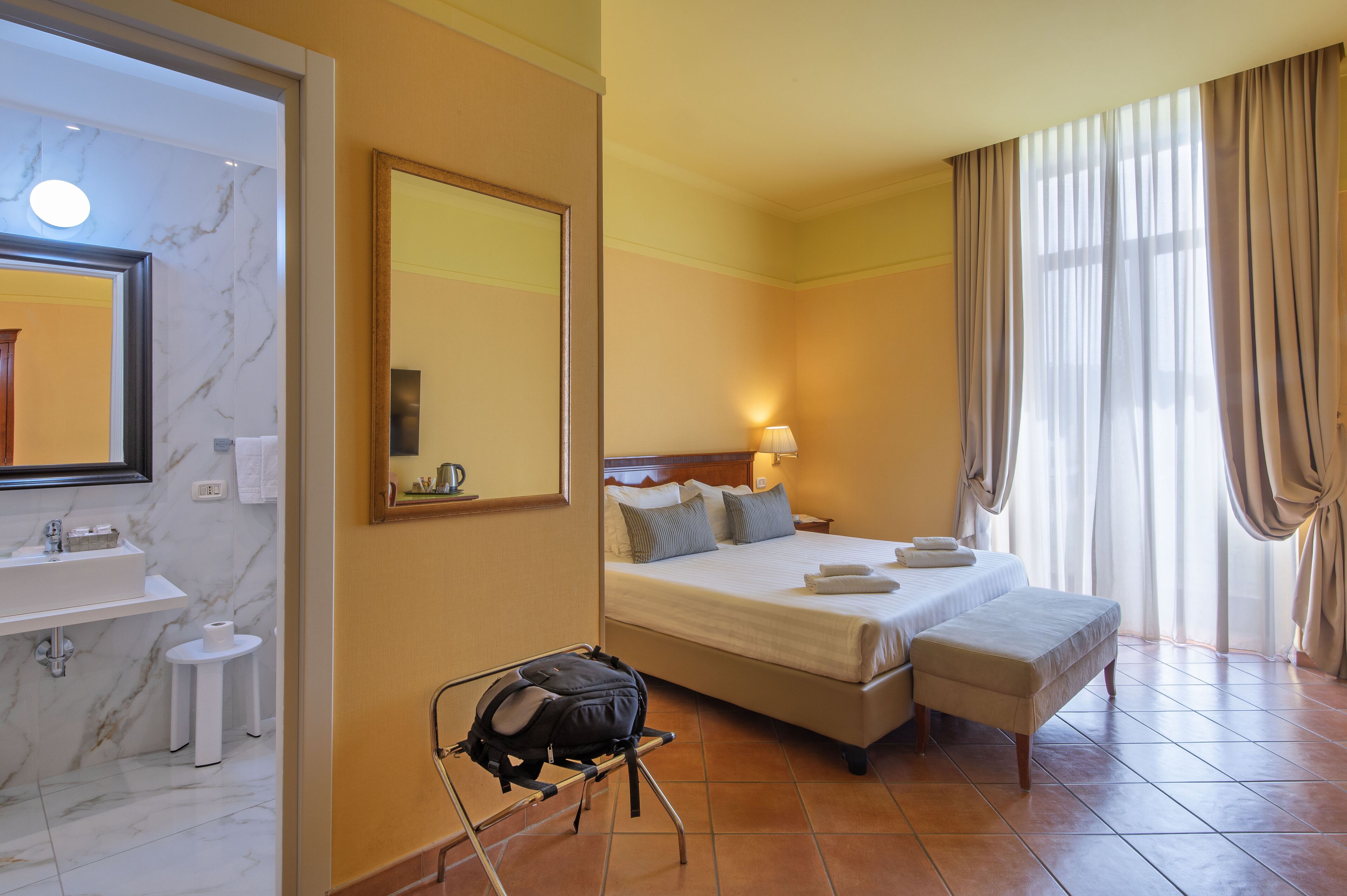 A hotel room with a marble bathroom, a bed with pillows, a bedside table, and a suitcase on a trolley.