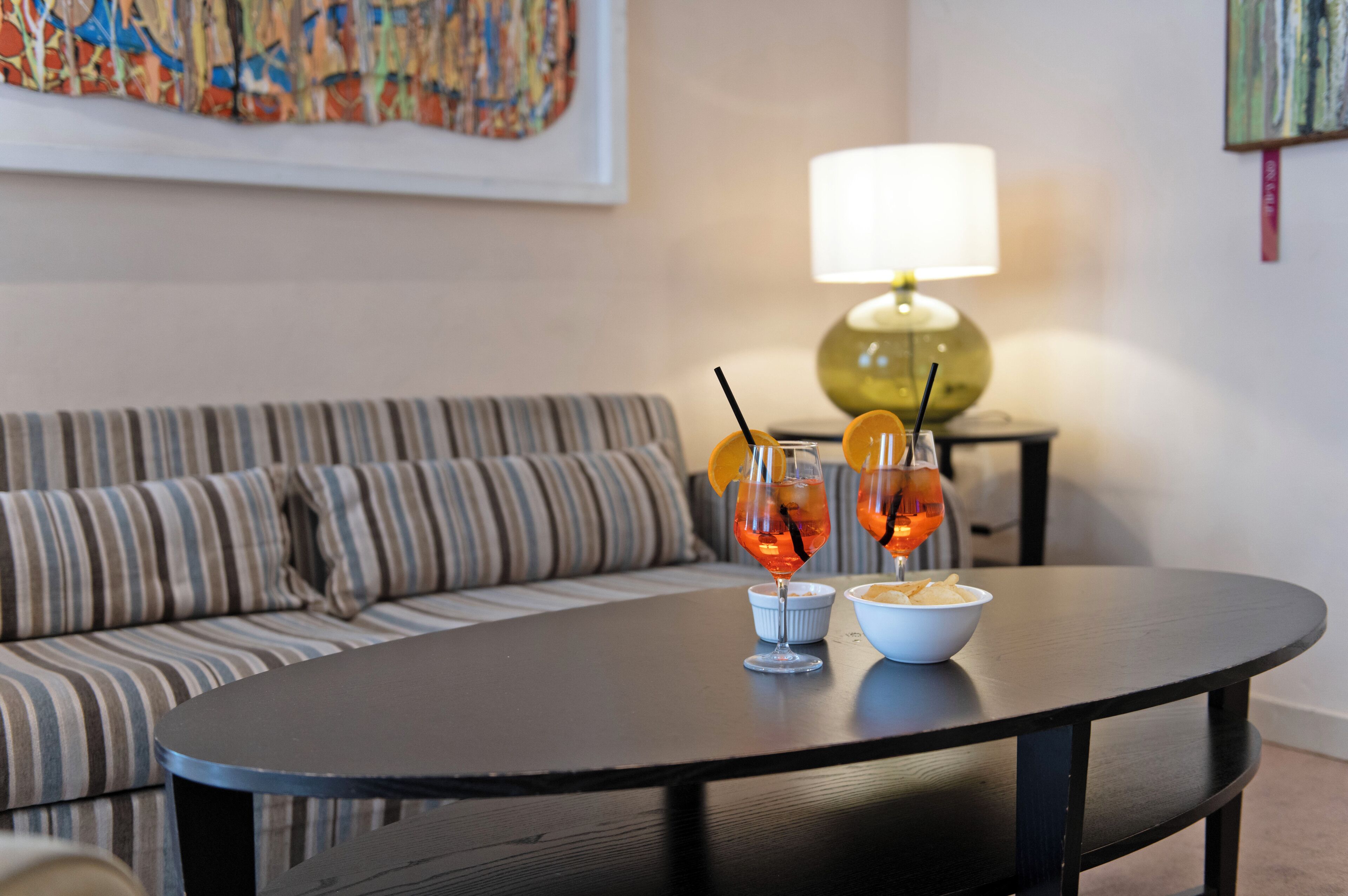 Ein modernes Wohnzimmer mit einem runden schwarzen Tisch, zwei Gläser Orangensaft, einer Schüssel Chips, einem gestreiften Sofa und einer Lampe auf einem Beistelltisch.