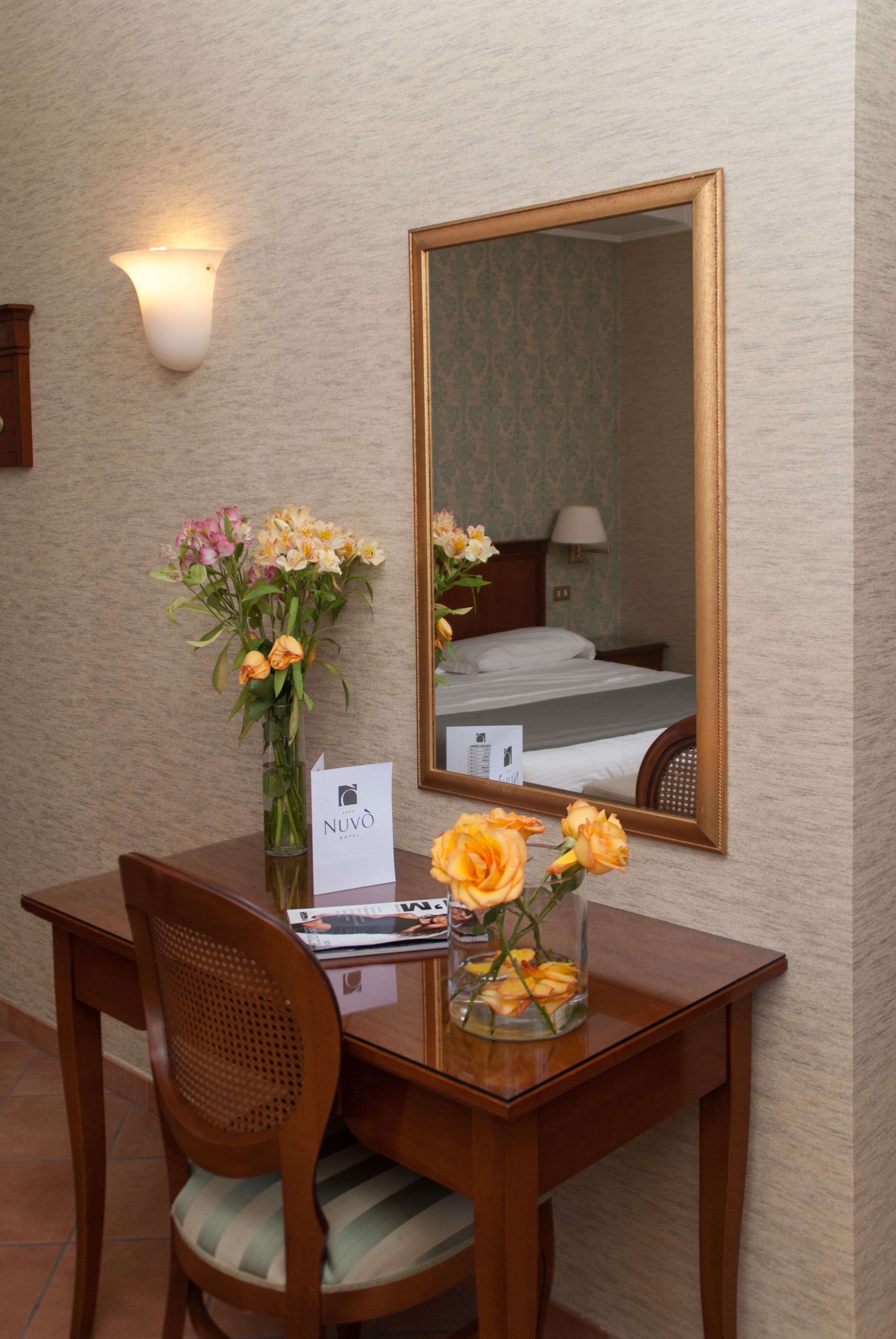 Habitación de hotel con escritorio de madera, una silla, un florero con flores, un espejo y una cama con mesita de noche.