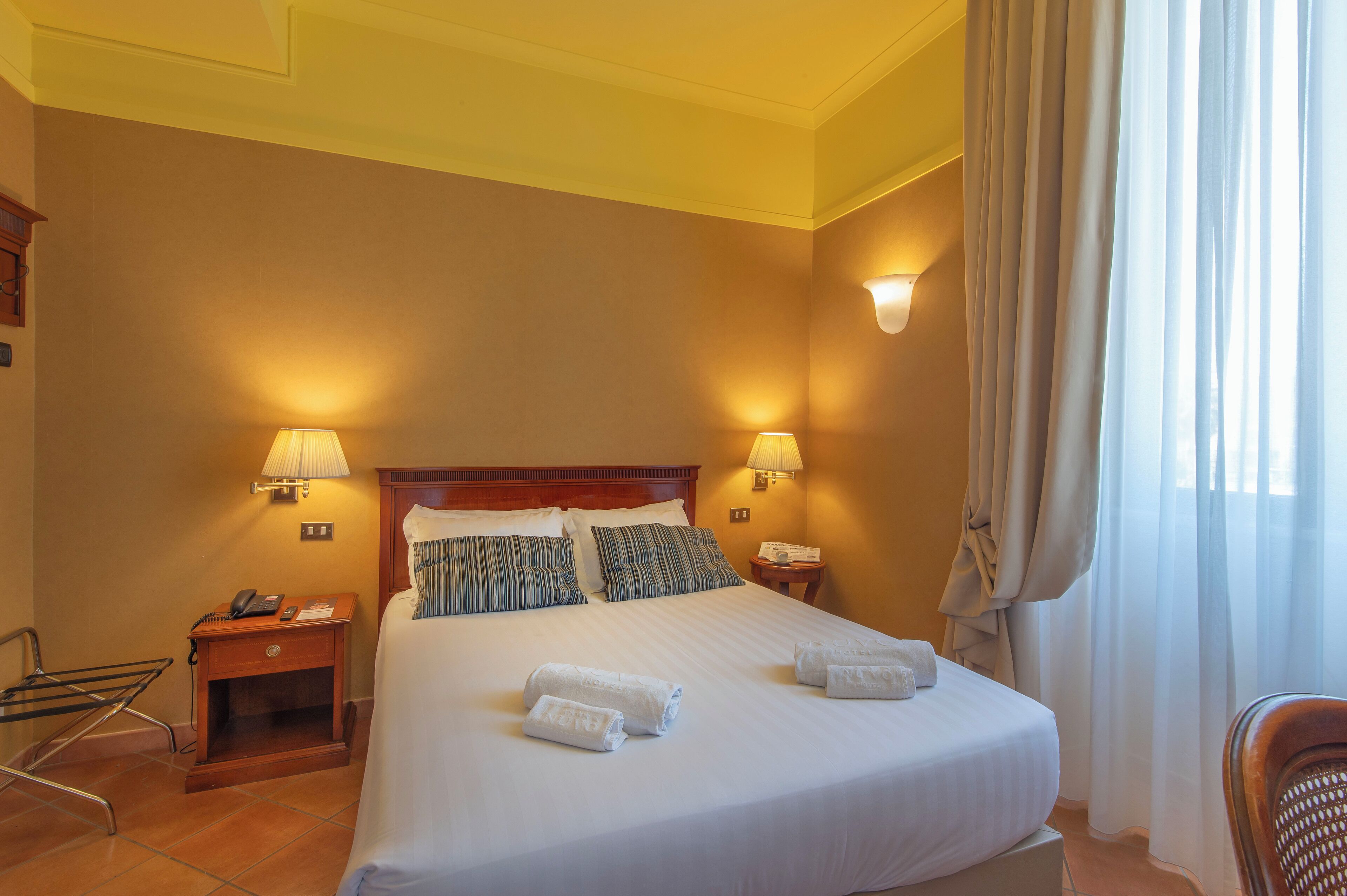 A hotel room with a large bed, two bedside lamps, a wooden nightstand, a chair, and a window with curtains.